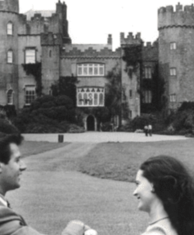 A black-and-white photo of a man and woman smiling and holding hands in the foreground, with a historic, castle-like building in the background. The castle has turrets and a large entrance, and is surrounded by a lawn and pathways.