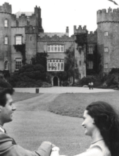 A black-and-white photo of a man and woman smiling and holding hands in the foreground, with a historic, castle-like building in the background. The castle has turrets and a large entrance, and is surrounded by a lawn and pathways.