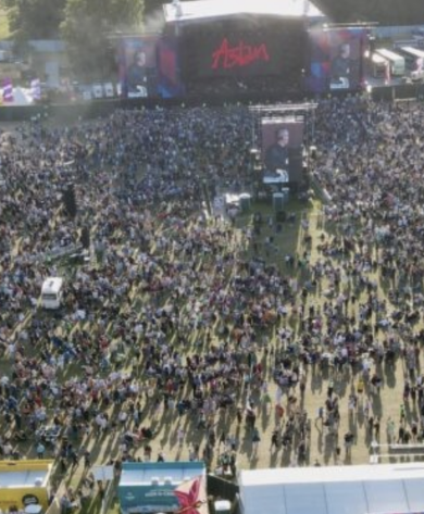 A large crowd of people attending an outdoor music festival during the day. The stage at the front features a red word "Asian" on a black background with screens displaying performers. Tents and vendor booths are visible around the perimeter.