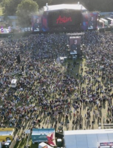 A large crowd of people attending an outdoor music festival during the day. The stage at the front features a red word "Asian" on a black background with screens displaying performers. Tents and vendor booths are visible around the perimeter.