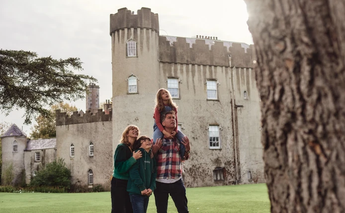 A joyful family of four stands in front of a historic castle. A woman and two children, one on the man's shoulders, laugh and look up in awe at the tree in front of them. They stand on a grassy lawn with the castle’s stone facade and turrets as the backdrop.