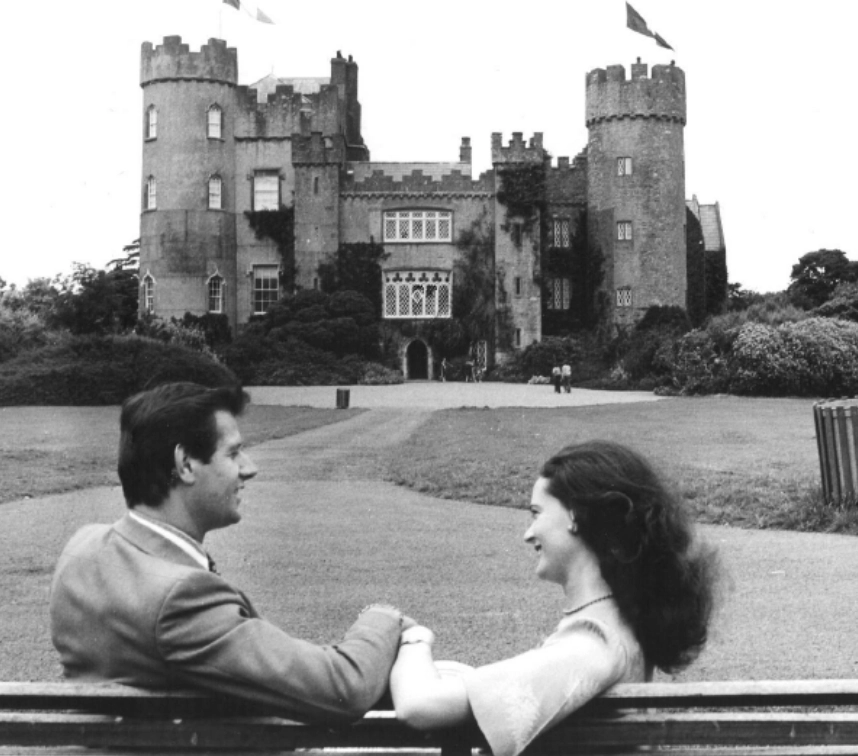 A man and woman sit closely on a bench, smiling at each other in front of a historic castle with towers and battlements. The castle has arched windows and is surrounded by greenery and a spacious lawn.