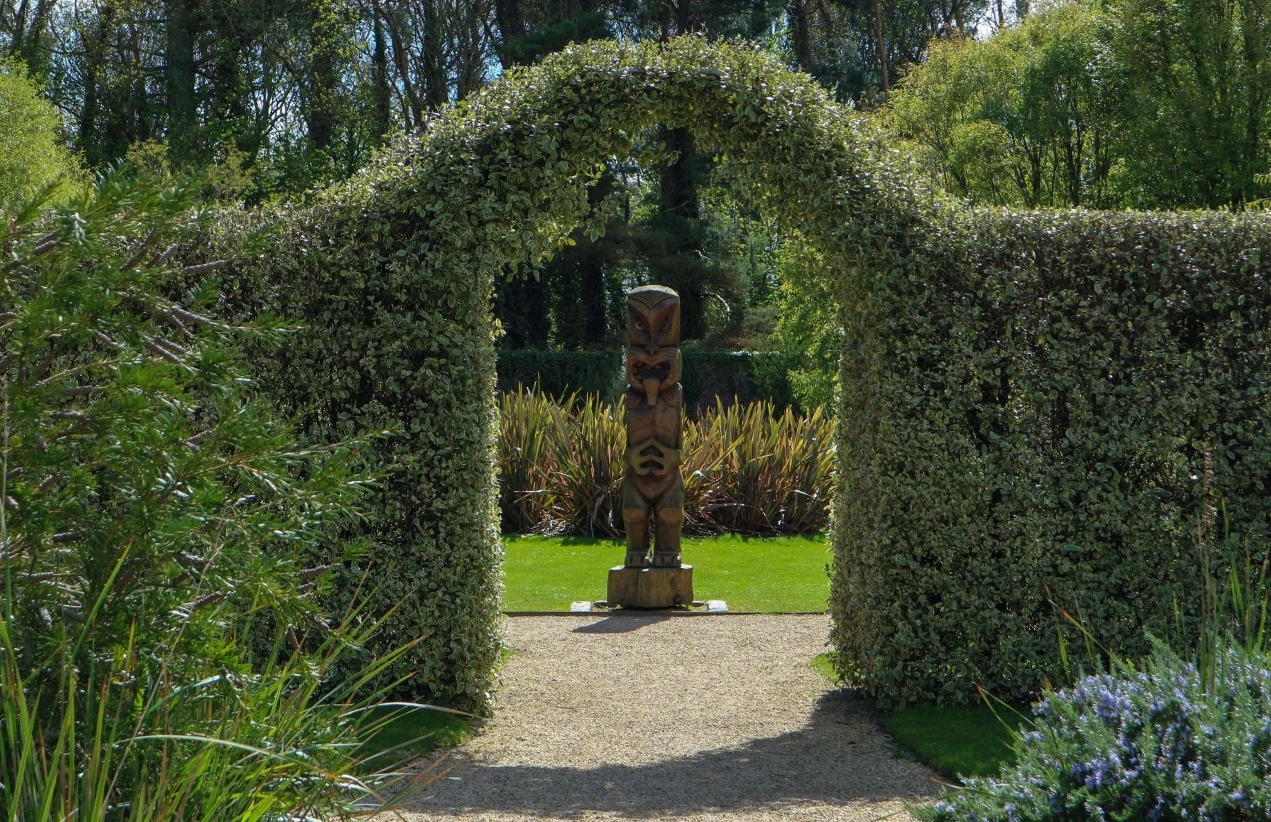 A lush garden at Malahide Castle features a tall wooden totem pole standing on a gravel path, framed by an archway of dense green foliage. Sunlight filters through the trees, highlighting the vibrant greenery and landscaped surroundings.