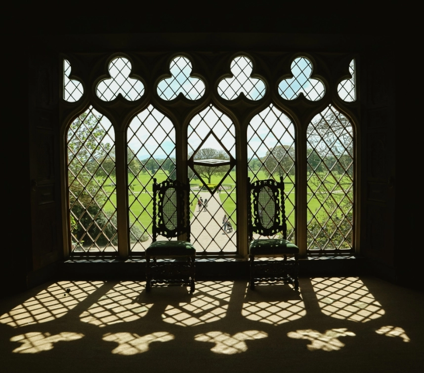 Two intricately carved chairs sit in front of a large, multi-paned Gothic-style window, casting elaborate lattice shadows on the floor. The window overlooks a bright, grassy landscape with a tree-lined path in the distance under a blue sky.