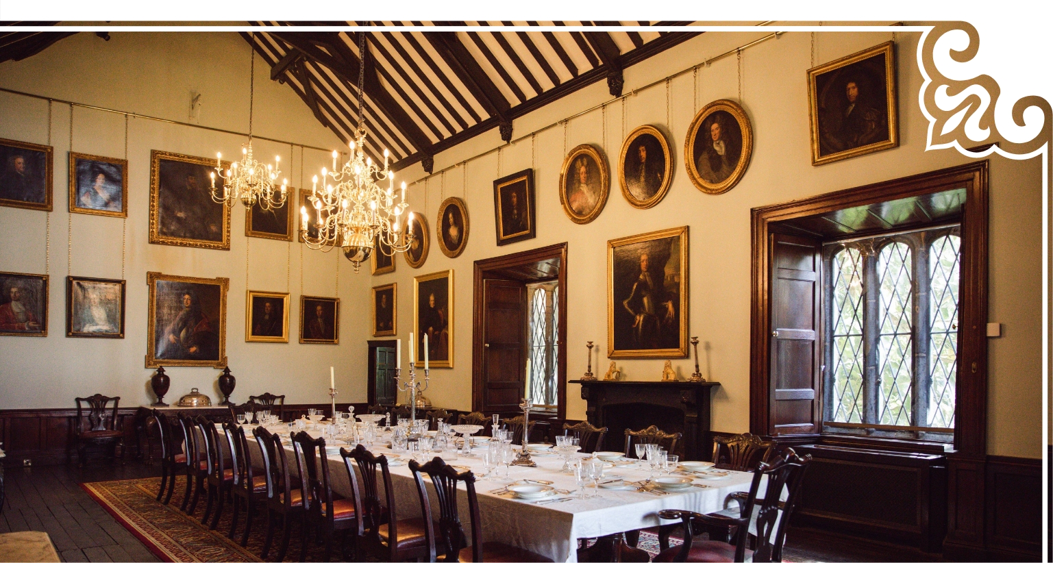 A grand dining room in Malahide Castle features a long table set for a formal meal. Chandeliers hang from the high, beamed ceiling, while portraits in ornate frames adorn the walls. Tall, arched windows let in natural light.