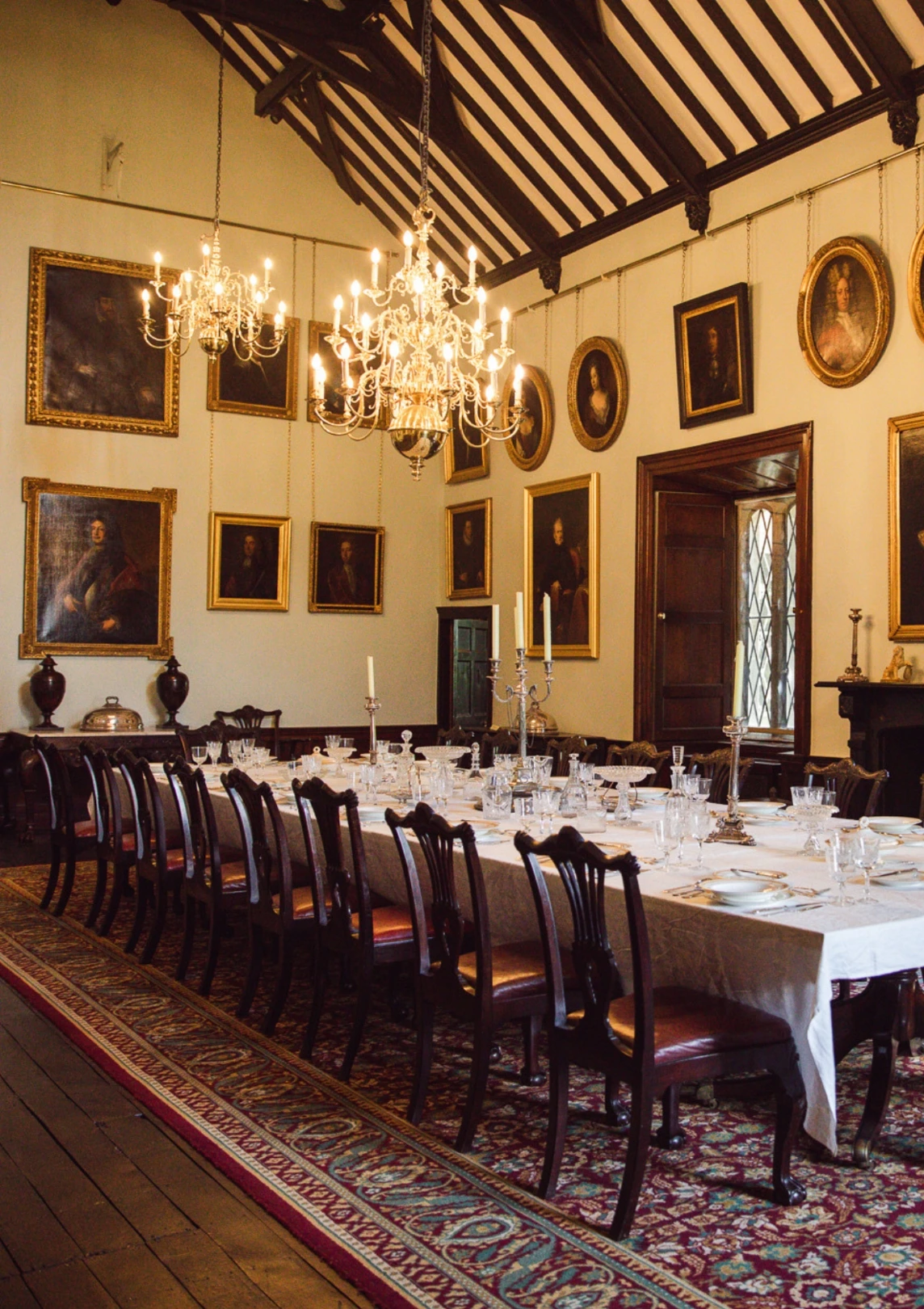 A grand, ornate dining room reminiscent of Malahide Castle, with a long wooden table set for a formal dinner. The room features high, beamed ceilings, two large chandeliers, numerous portrait paintings on the walls, and intricate rug flooring. The table is adorned with fine china and candlesticks.